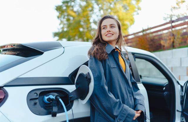 Mulher jovem satisfeita aguardando o seu carro elétrico carregar - Adobe